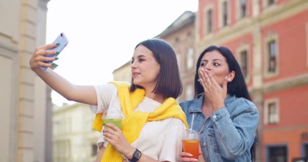 Mulheres muito caucasianas se divertindo tirando fotos, mexendo, sorrindo. Jovens amigas segurando bebidas frescas, tomando selfie no telefone, soprando beijos ao ar livre. Amizade, conceito de tecnologia. — Vídeo de Stock