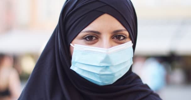 Potrairt of pretty muslim woman wearing hijab, hiding her face outdoors on crowded street. Adult female arab using medical mask to protect from coronavirus, looking to camera and smiling. Health. — Stock Video