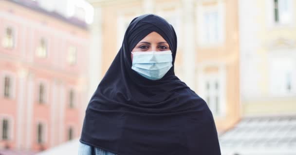 Atraente velado muçulmano fêmea em pé na rua e olhando para a câmera. Mulher árabe adulto em máscara protetora médica vestindo hijab ao ar livre. Religin, pessoas, conceito de cultura. — Vídeo de Stock