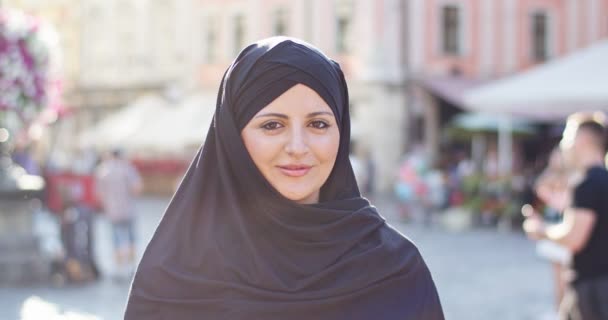 Retrato de una mujer árabe velada y atractiva caminando por la calle de la ciudad en un día soleado. Joven musulmana vestida con hiyab negro, cubriendo la cabeza, mirando a la cámara y sonriendo. Religión, concepto de derechos. — Vídeos de Stock