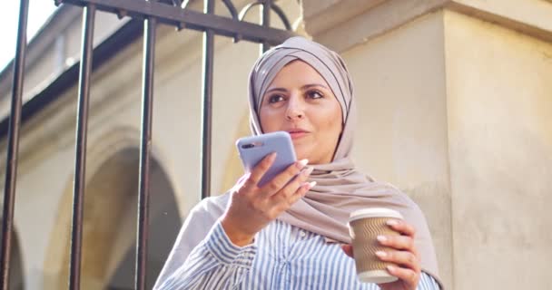 スマートフォンを使用して魅力的な女性のアラブ人は、屋外で友人の家族とチャット。若いイスラム教徒の女性は、録音、音声メッセージを送信ヒジャーブを着てコーヒーカップを保持。技術、コミュニケーション. — ストック動画