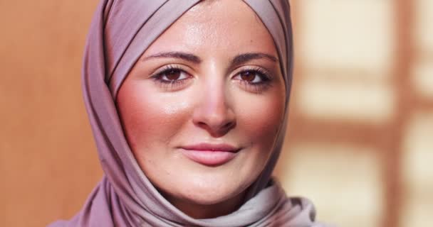 空の日当たりの良い部屋に立ってベール頭を持つ美しい女性のアラブ。大人のイスラム教徒の女性をカバーするために紫色のヒジャーブを着て、カメラと笑顔を探しています。宗教、美の概念. — ストック動画