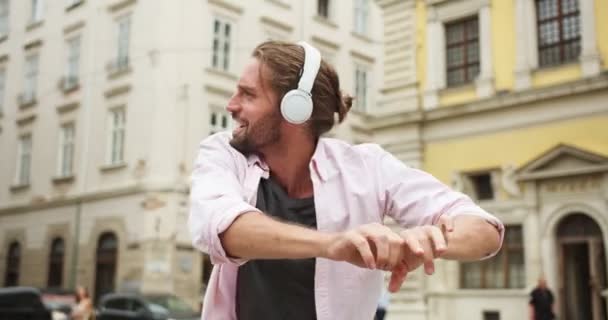 Çekici beyaz adam dans ediyor, etrafta takılıyor, sokaklarda takılıyor. Kulaklıklı genç adam akıllı telefondan müzik dinliyor, MP3 uygulamadan. Eğlence, teknoloji konsepti. — Stok video