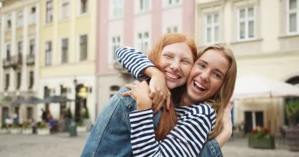 Två unga kvinnor som kramas, kramas, tittar på kameran på gatan i stan. Ganska kaukasiska kvinnliga bästa vänner myser och skrattar. Vänskap, förhållande, kvinnlighet. — Stockvideo