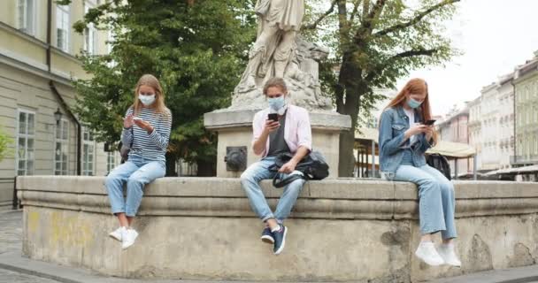 Tres jóvenes estudiantes masculinos y femeninos sentados en el banco de la fuente y usando teléfonos inteligentes. Gadget adicto caucásico hombre y mujeres mensajes de texto, no hablar entre sí. Redes sociales. — Vídeo de stock