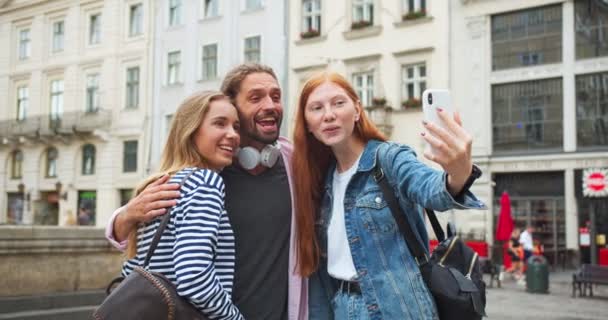 Kaukázusi fiatalok fotóznak és videóznak okostelefonon a városban. Jóképű lányok és a srác a kamerába tekergőznek és nevetnek a szabadban. Technológia, ifjúság, szórakozás. — Stock videók