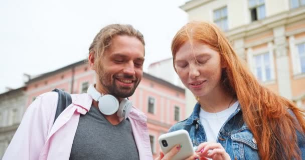 Attraktiv rödhårig kvinna som rekommenderar sång till sin manlige vän. Ung kille njuter av att lyssna på musik tänd av dam på smartphone i stan. Underhållning, teknik, app-koncept. — Stockvideo