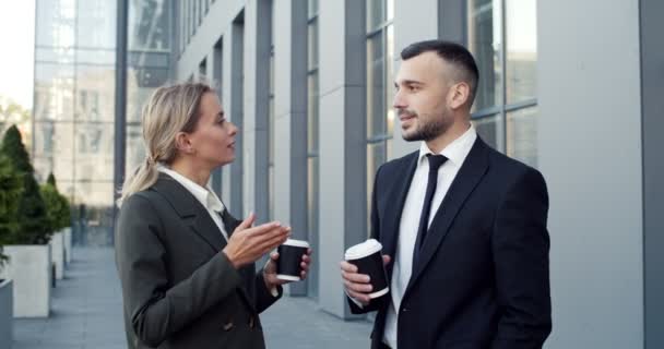 Baik mencari rekan bisnis laki-laki dan perempuan muda berdiri di jalan dekat gedung kantor besar, berbicara satu sama lain dan menyesap kopi panas untuk-pergi — Stok Video