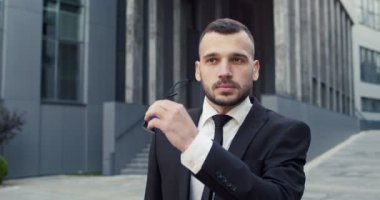 Yakışıklı genç girişimci güneş gözlüklerini çıkarıyor ve şehirde sokakta tek başına dikilirken kameraya bakıyor. Kafkas erkek yöneticisinin yakın portresi. İş adamı, akıllı görünüş.