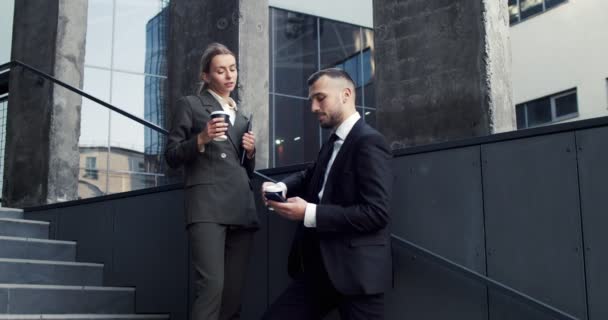 Bons jeunes collègues d'affaires masculins et féminins qui descendent du grand immeuble de bureaux, se parlent et sirotent du café chaud à emporter — Video