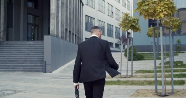 Zadní pohled na zaneprázdněného manažera mužského pohlaví držícího smartphone při běhu venku v blízkosti business centra. Bělošský podnikatel si pospíší na ulici — Stock video