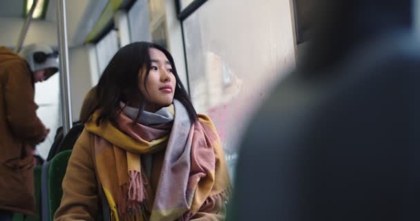 Attractive stylish Asian young girl smiling and looking at the window while going somewhere and sitting in the tram or bus. — Stockvideo