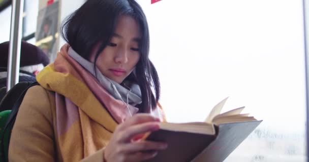 Asian beautiful young woman in headphones sitting at the window at the tram or bus and reading interesting funny story in a book so smiling. Close up. — Stockvideo
