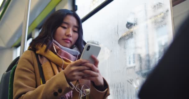 Joven mujer bonita asiática en auriculares escuchando la música y grabando en el teléfono inteligente mientras se sienta en la ventana y va a algún lugar en el tranvía o autobús. — Vídeo de stock