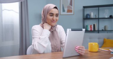 Güzel tesettürlü mutlu Müslüman bir kadının tabletle görüntülü konuşma yapıp modern bir odada oturup arkadaşlarına gülümsemesi. Yaşam tarzı, modern İslam kavramı.