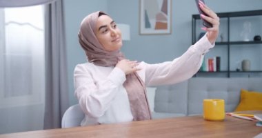 Genç müslüman bir kadının modern iç mekanda selfie çektiği bir plan. Lüks evde kamera arayan tesettürlü çekici bir kadın. Evde masa başında kameraya poz veren oyuncu kadın..