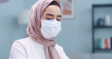 Profesyonel müslüman iş kadınının yüz maskesi takıp kameraya bakan portresi. Ev ofisinde geleneksel başörtüsü takan Arap bir kadın. Uzak çalışma, çevrimiçi konsept eğitimi.