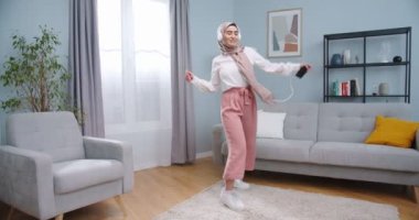 Şakacı, tesettürlü, beyaz kulaklıklı, akıllı telefondan müzik dinleyen ve modern tarz oturma odasında dans eden komik Arap kadın. Kadın evde kalırken eğleniyor..
