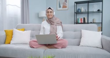 Kafiyeli neşeli Müslüman bir kadının orta planı. Bilgisayardan müzik dinliyor, kulaklık takıyor, dans ediyor, kafiye yapıyor. Güzel, minimalist oturma odasında eğlenen bir kadın..