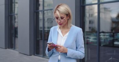 Beyaz tenli, gözlüklü güzel bir kadın. Ofis çalışanı. Elinde akıllı telefonla konuşuyor. Modern binanın önünde durup mesajlaşıyor ve gülümsüyor..