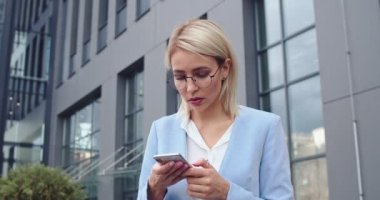 Şık ve yakışıklı beyaz iş kadını akıllı telefondan bir şeyler izliyor, gülümsüyor ya da sosyal medyadaki mesajlarla sohbet ediyor. Dışarıda..