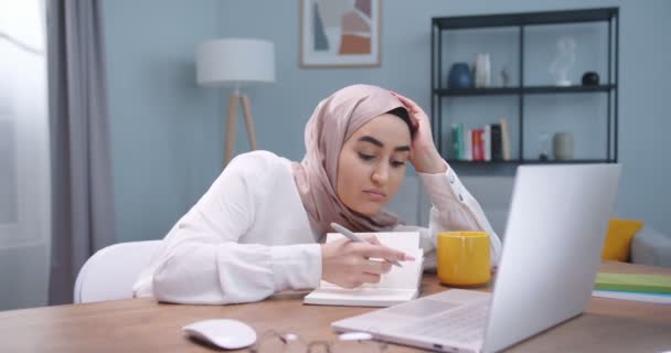 Plano medio de cansado estudiante musulmán agotado usando hijab, estudiando duro en el escritorio en la habitación, usando computadora portátil y cuadernos, preparándose para los exámenes. Estilo de vida de los estudiantes, concepto islam moderno — Vídeos de Stock