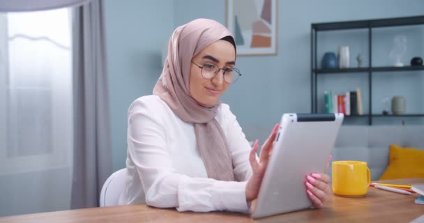 Middle plan of happy muslim woman in hijab and glasses sitting at cozy room, using digital tablet and smiling, serfing, browsing in Internet. Joyful female texting friend online. Concept of technology — Stockvideo