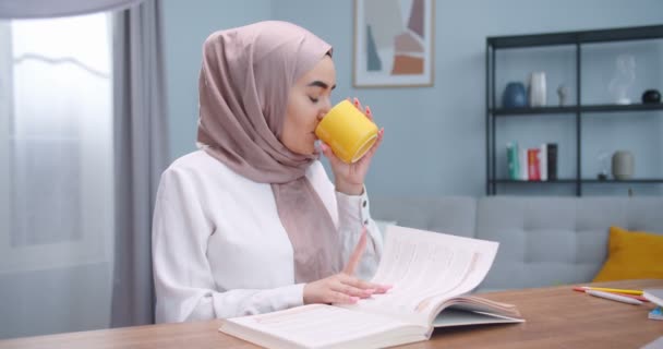 Middle plan of muslim young prettywoman in hijab reading an interesting story in book at home in the cozy room and drinking tea from yellow cup. Smart serious student stadying, prepearing for exams. — Stock Video