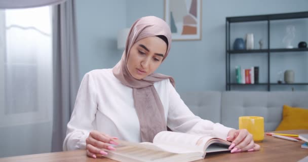 Środkowy plan muzułmanki czytającej książkę, siedzącej przy biurku w nowoczesnym, stylowym salonie. Kobieta studiująca literaturę edukacyjną. Kształcenie na odległość. Styl życia studentów, nowoczesna koncepcja islamu — Wideo stockowe