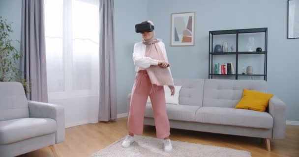 Joyeuse jeune femme arabe en hijab portant un casque de réalité virtuelle et tenant des contrôleurs jouant dans un jeu vidéo à la maison. Femme jouant VR tennis au milieu du salon. — Video