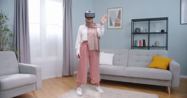Μεσαίο σχέδιο της όμορφης μουσουλμάνα γυναίκα σε μαντίλα φορώντας ακουστικά εικονικής πραγματικότητας και κρατώντας ελεγκτές κύλιση εικονική οθόνη στο σπίτι. Γυναίκα που διαμένει στο Google VR στη μέση του σαλονιού. — Αρχείο Βίντεο