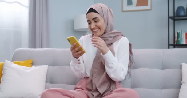 ヒジャーブ州の幸せなイスラム教徒の女性の中間計画は、自宅のオフィスで携帯電話を検索楽しむ。アパートの電話でインターネットでサーフィン驚くべき女性。楽しい女の子は電話で良いニュースを読んで. — ストック動画