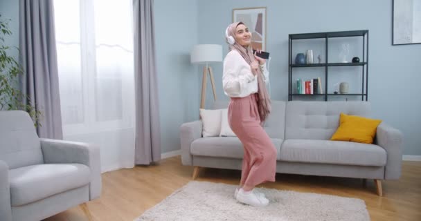 Дивовижна красива муслімна жінка в хіджабі слухає музику в білих головах з телефона і рухає своїм тілом в музичному ритмі, танцюючи в стильній вітальні. Аравійка добре бавиться, коли залишається вдома.. — стокове відео