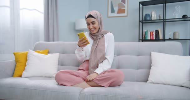 Plano medio de mujer musulmana relajada en hijab sosteniendo el teléfono inteligente mirando la pantalla riendo disfrutando usando aplicaciones móviles divirtiéndose jugando juegos charlando en redes sociales sentados en el sofá en casa. — Vídeo de stock