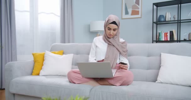 Plano médio da jovem mulher muçulmana feliz no hijab segurando cartão de crédito usando pagamentos móveis instantâneos em casa, comprador fazendo compra no telefone inteligente recebendo conceito de reembolso. Serviço de aplicativos de E-banking — Vídeo de Stock