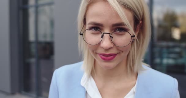 Portrait of the beautiful and happy blond Caucasian woman CEO in glasses looking down and then rising her sight to the camera and smiling. Outside. Close up. — 图库视频影像