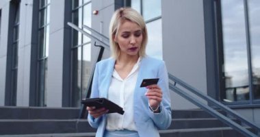 Beyaz tenli, iş stili çekici bir kadın elinde tablet bir cihaz tutuyor ve sokakta kredi kartıyla internetten alışveriş yapıyor..