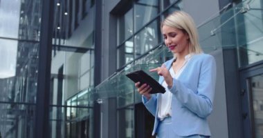 Beyaz saçlı, iş kıyafeti giymiş, modern ofis binasının dışındaki tablet bilgisayara tıklayıp daktilo eden ve kameraya neşeyle gülümseyen güzel beyaz kadının portresi..