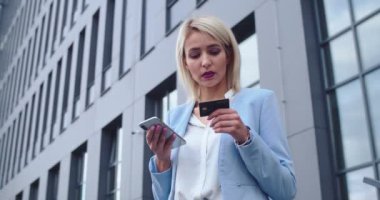 Sarışın, beyaz iş kadınının akıllı telefondan alışveriş yaptığı ve kredi kartıyla ödediği manzarayı izleyin. Açık havada..