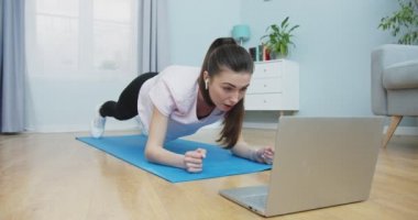 Kendine güvenen esmer spor giyimli bir kadının orta planı, bilgisayar kullanarak kulaklıklarda konuşma ve dinleme yaparken tahta egzersizleri yapmaktır. İnsanlar ve aktif yaşam tarzı kavramı