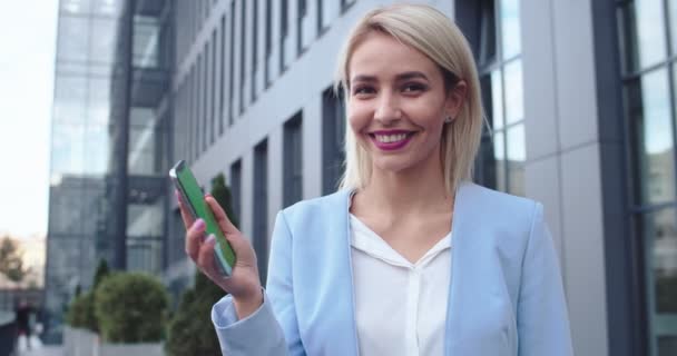 Portrait shot of the beautiful and cheerful blonde businesswoman standing outdoors with a smartphone in hand, then demonstrating phone with a green screen to the camera. Chroma key. Tracking motion. — Vídeo de Stock