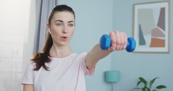 Close up of sporty woman in white headsets working out with dumbbells in living room while watching online training. Focused and concentrated woman training her arms. Sport and fitness. — Stock Video
