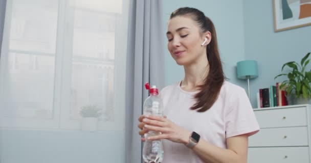Porträtt av bedårande ung och vältränad kvinna sitter på golvet hemma efter intensiv sport träning och dricksvatten från flaska och tittar till kameran. Träning, träning och wellness koncept. — Stockvideo