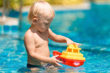 Küçük sarışın bir çocuk, çocuk havuzunda bir tekneyle oynuyor.