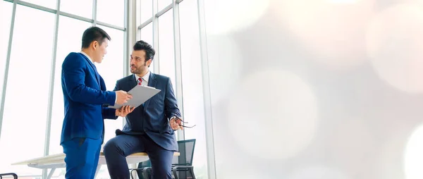 Two business people talk project strategy at office in widen view meeting room. Businessman discuss project planning with colleague at modern workplace .