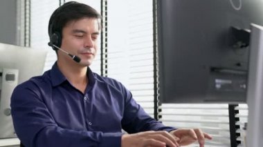 Young competent asian male call center agent working at his computer while simultaneously speaking with customers. Concept of an operator, customer service agent working in the office with headset.