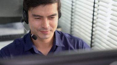 Young competent asian male call center agent working at his computer while simultaneously speaking with customers. Concept of an operator, customer service agent working in the office with headset.