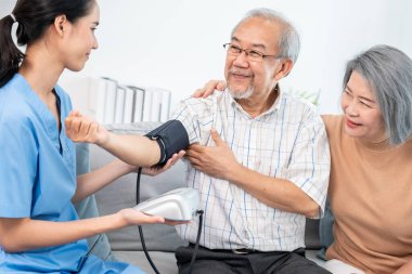 An elderly man having a blood pressure check by his personal caregiver with his wife sitting next to him in their home.