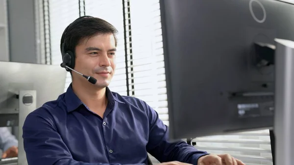 Young competent asian male call center agent working at his computer while simultaneously speaking with customers. Concept of an operator, customer service agent working in the office with headset.