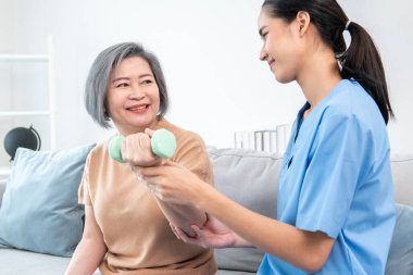 Caregiver helping contented senior woman exercise with dumbbell at home. Rehab for elder, senior medical care.